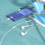 Smartphone playing music on a teal surface with white wired earphones resting nearby and a charging cable connected.